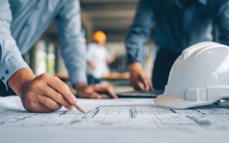 Construction workers are collaborating on detailed architectural plans at a building site. A hard hat is placed on the table as they mark key sections with pencils