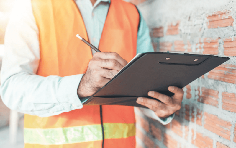 Mann in Warnweste schreibt auf Klemmbrett neben Ziegelwand.